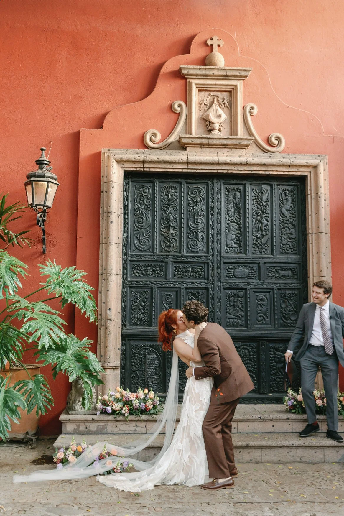 Newlywed couple kissing in front of an ornate black door on a terracotta building, with wedding flowers and guests cheering them on