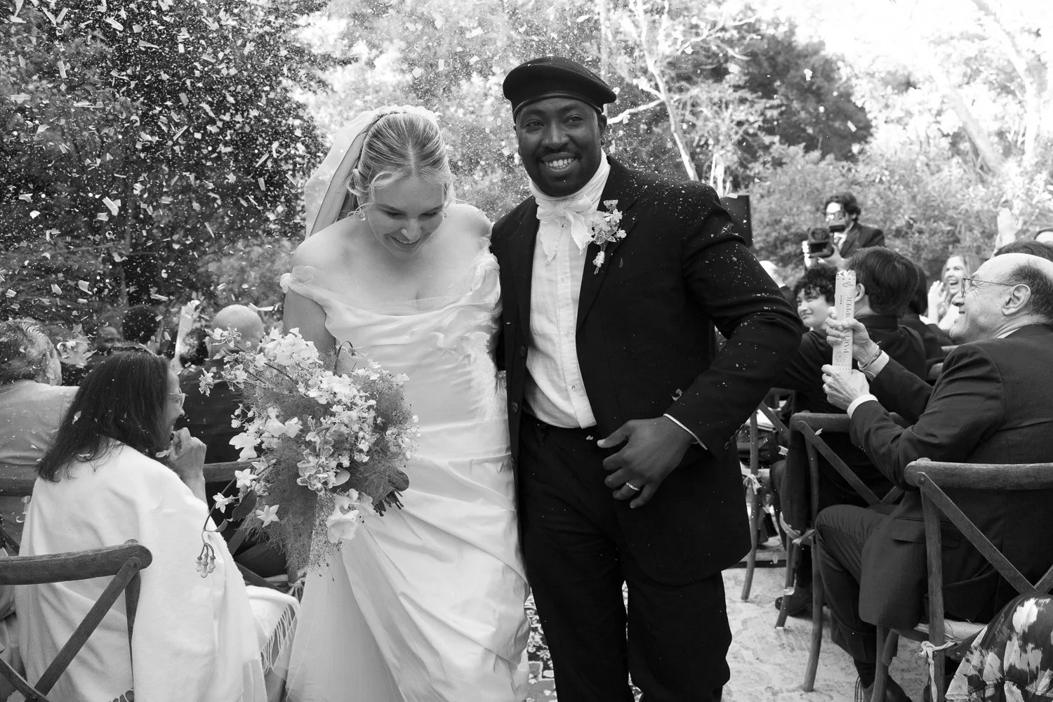 Black and white photo of a wedding celebration. A bride in a wedding dress and a groom in a tuxedo are smiling, with guests around them clapping and celebrating outdoors.