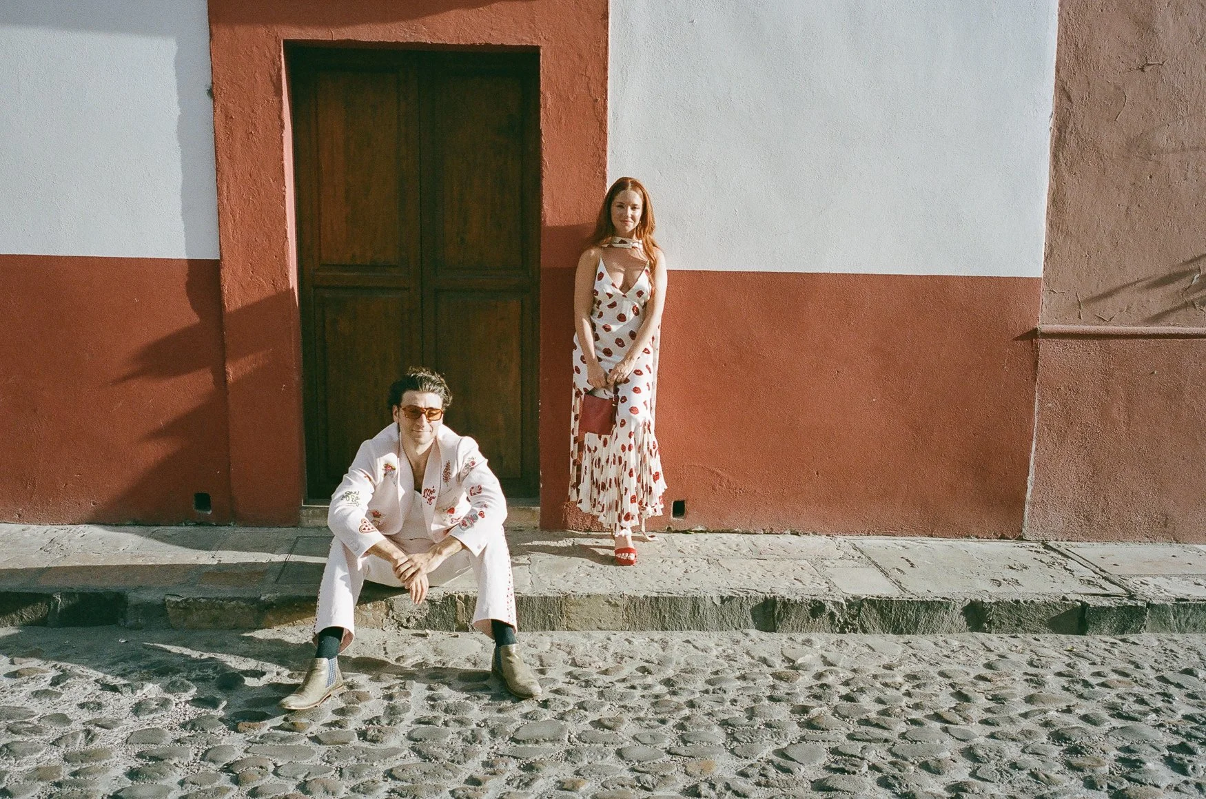 Two fashionably dressed people posing outside a colorful stucco building on a cobblestone street in san miguel de allende
