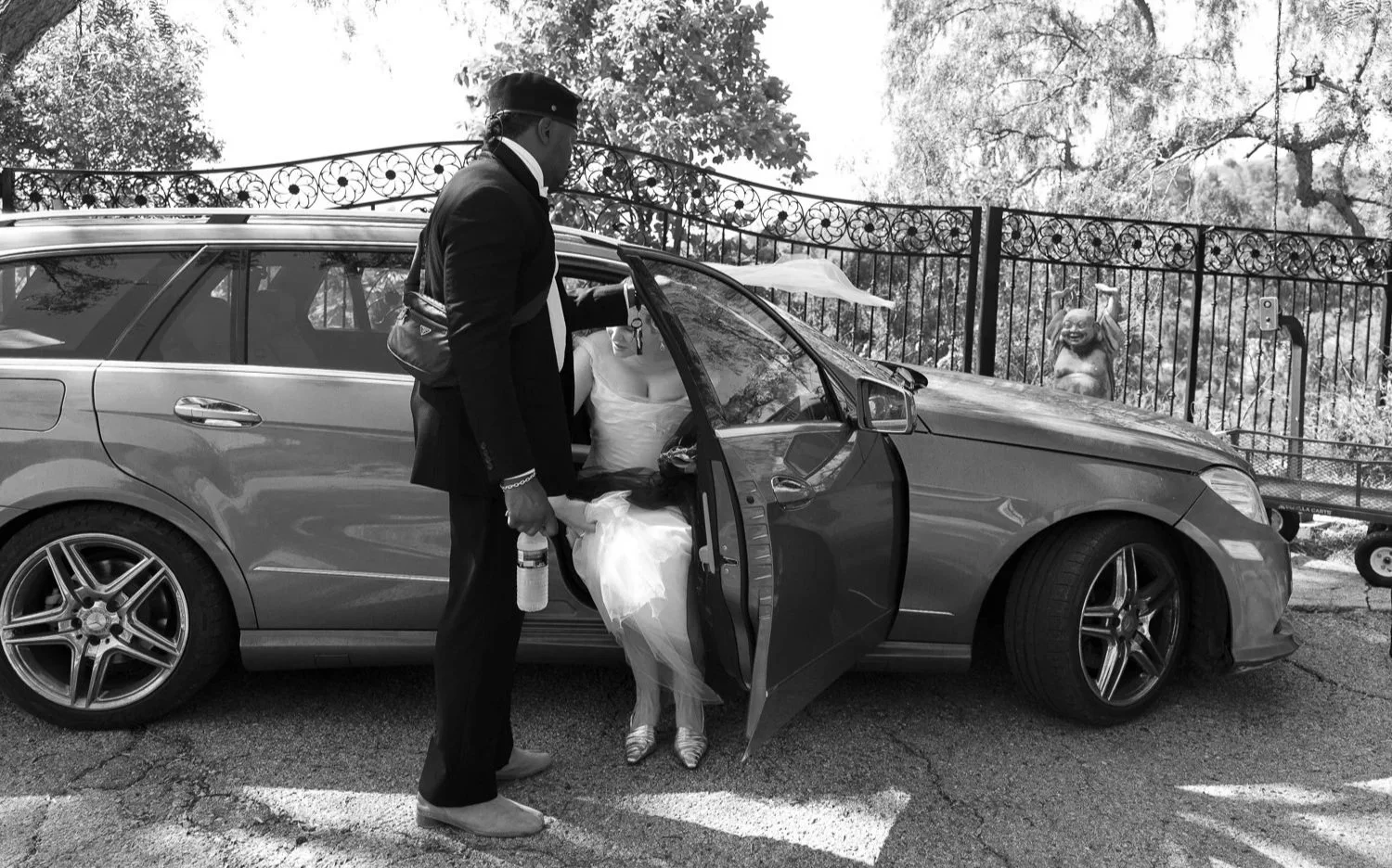 A man in a formal suit stands outside a car, holding the door open for a person in a wedding dress entering the vehicle, with a protected outdoor area with a fence and a decorative statue in the background.