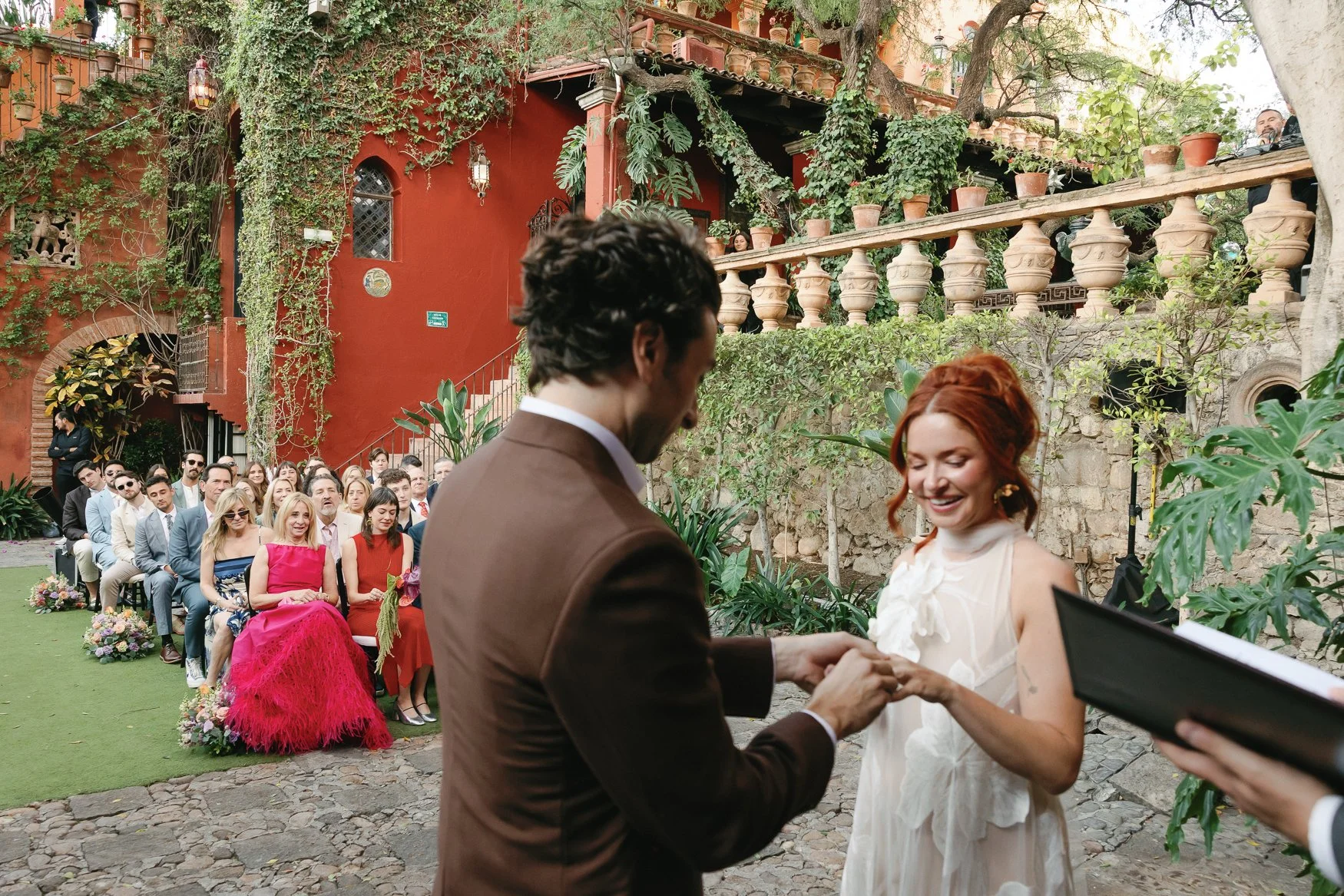 Bride and groom exchange rings at an outdoor wedding ceremony surrounded by seated guests and lush greenery.