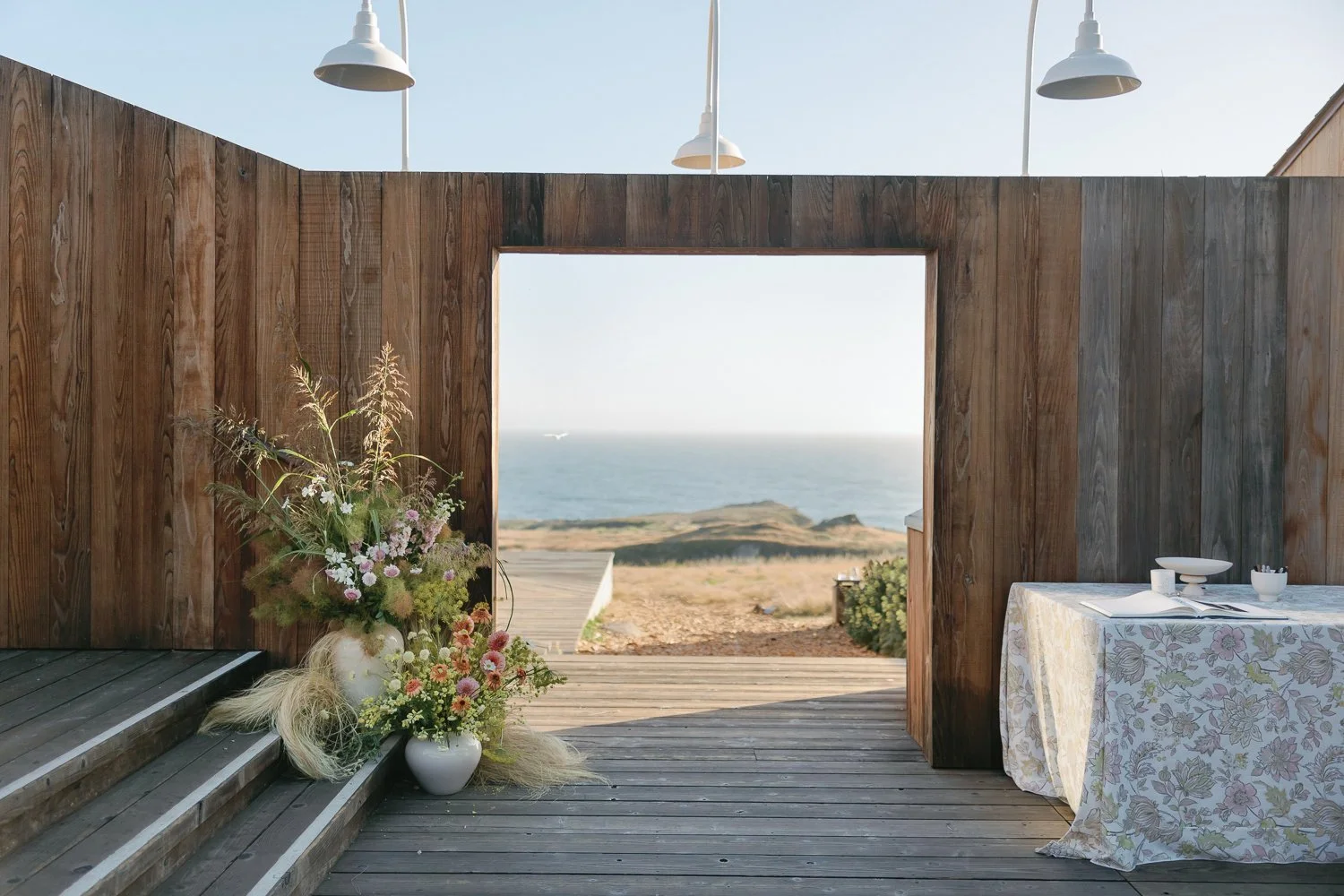 A wooden patio with a view of the ocean, a large flower arrangement in a white vase, and a table with a floral tablecloth and dishware.
