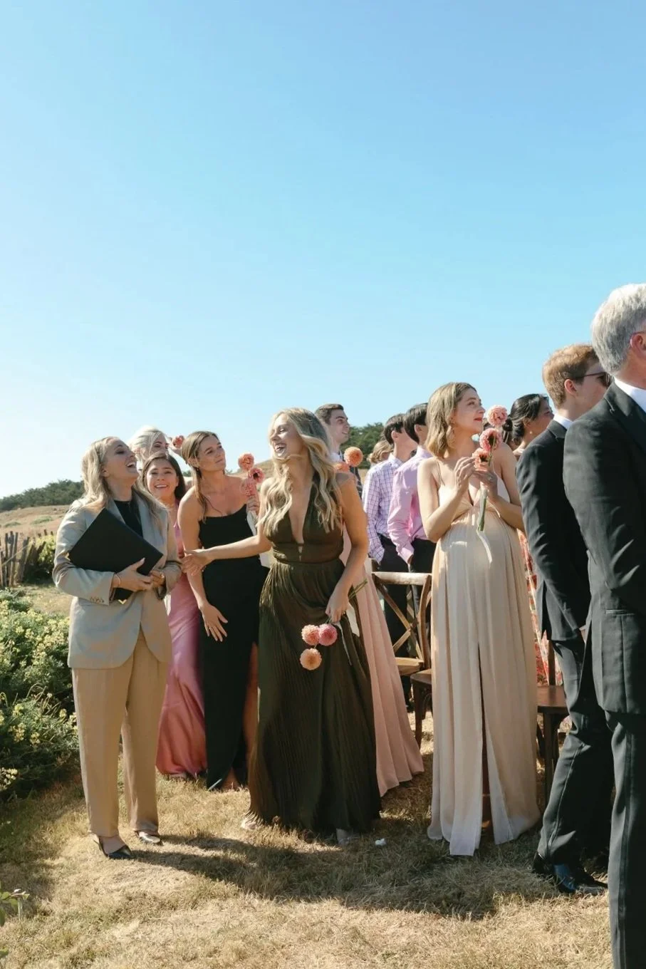 People dressed in formal attire attending an outdoor wedding ceremony, some holding pink flowers, under a clear blue sky with a scenic landscape in the background.