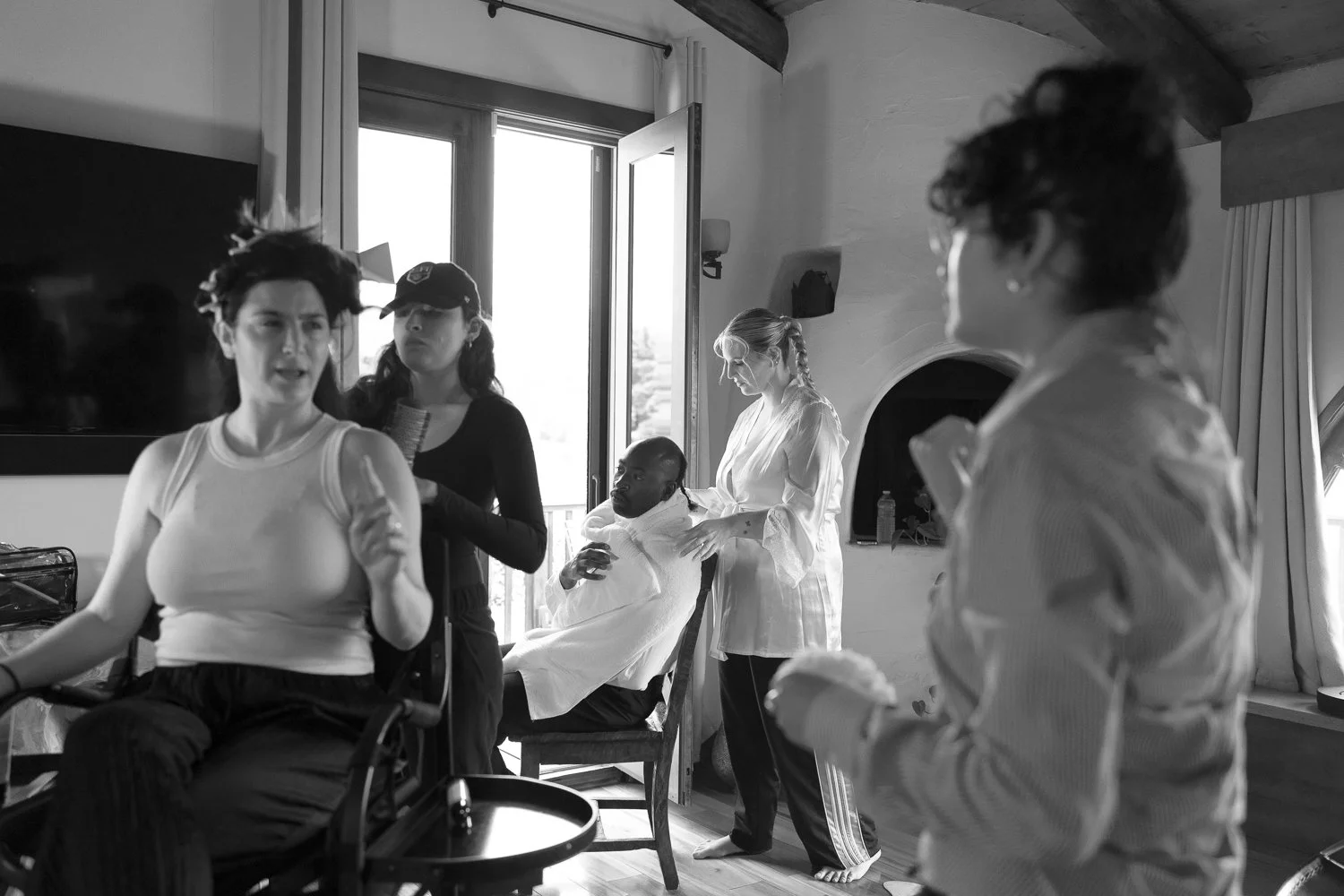 A black and white photo of a group of women in a room, engaging in discussions or preparations. One woman is seated in a chair with a white gown, possibly getting ready for a special occasion, while others stand around her, some with hair or makeup tools. A woman in the foreground appears to be talking or explaining something, and there is a woman with glasses and curly hair visible on the right.