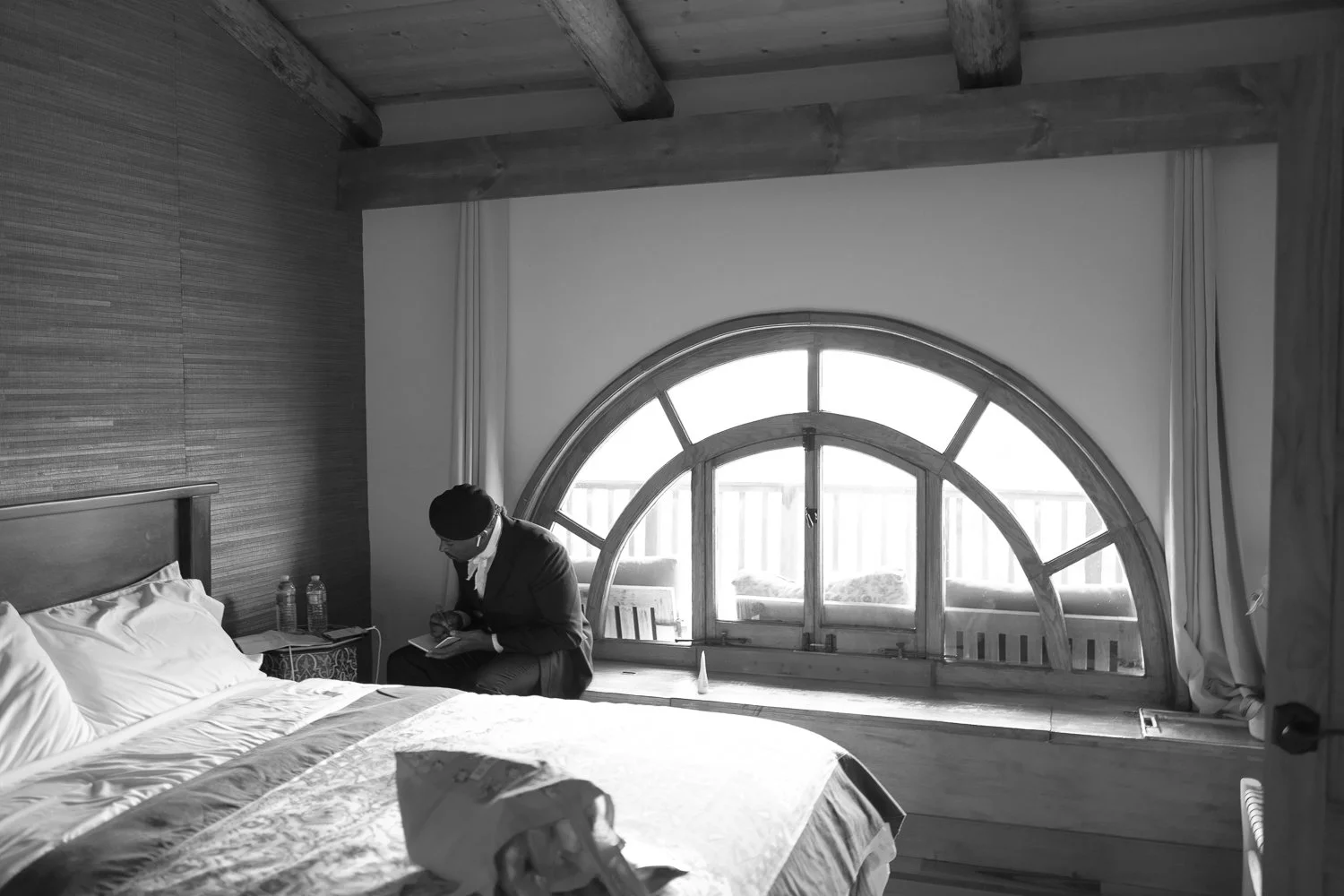 A man sitting on the edge of a bed writing his vows in a bedroom with a large semi-circular window and wooden ceiling beams.