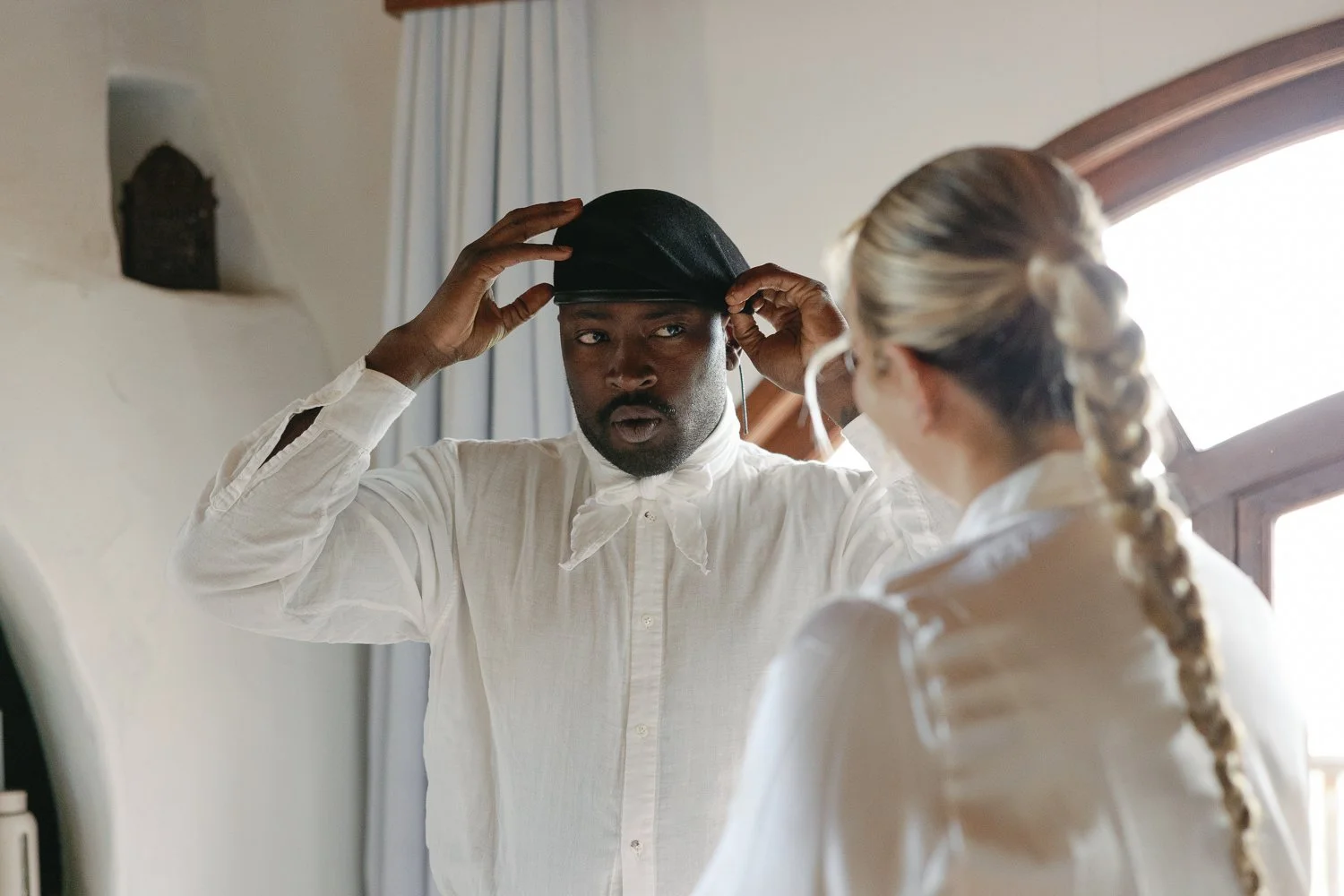 A man adjusting a black hat on his head while facing a woman with blonde hair tied in a braid, inside a room with a window and light-colored walls.