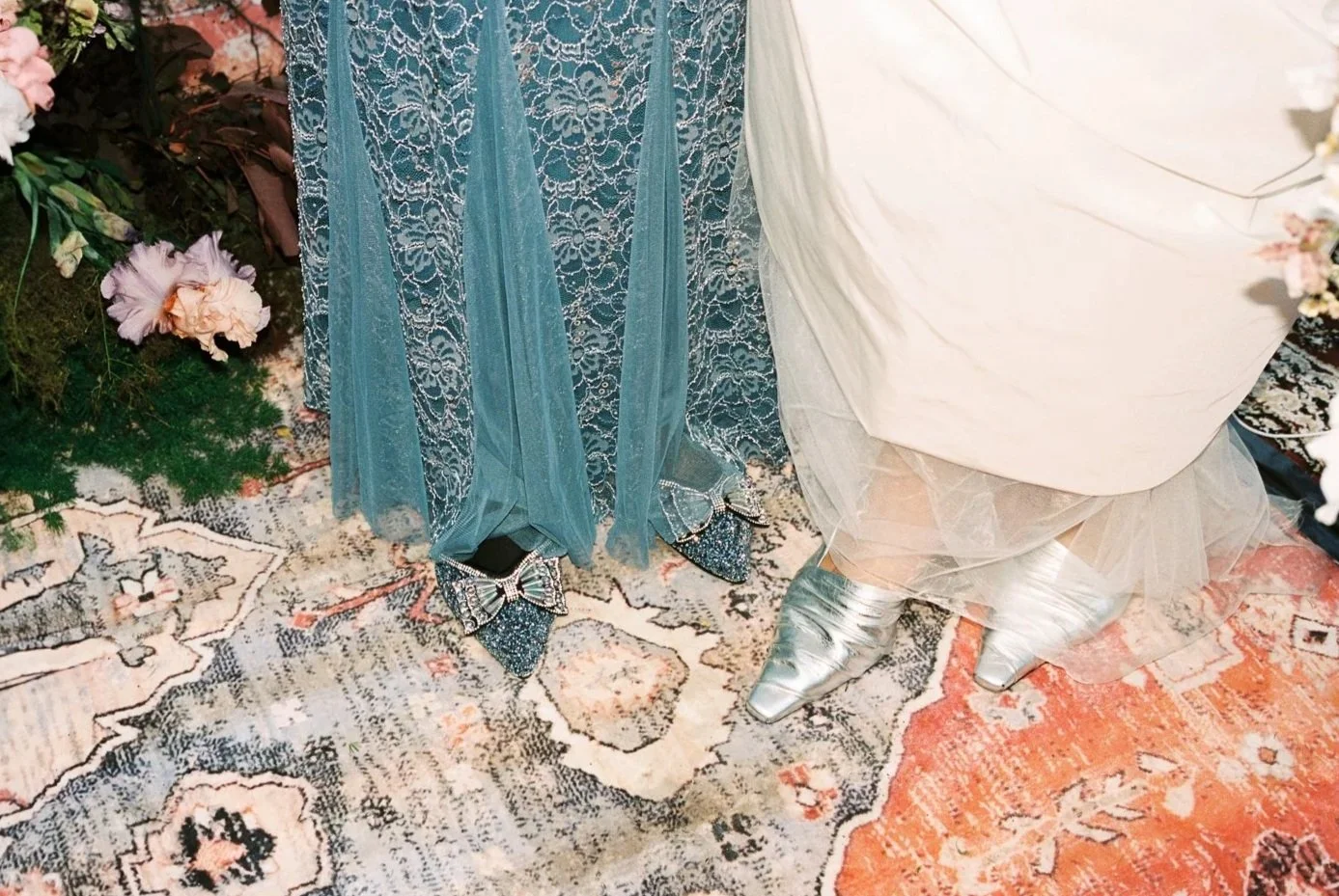 Part of a wedding dress, blue lace and tulle fabric, with sparkling silver shoes, surrounded by floral decorations and an ornate patterned rug.