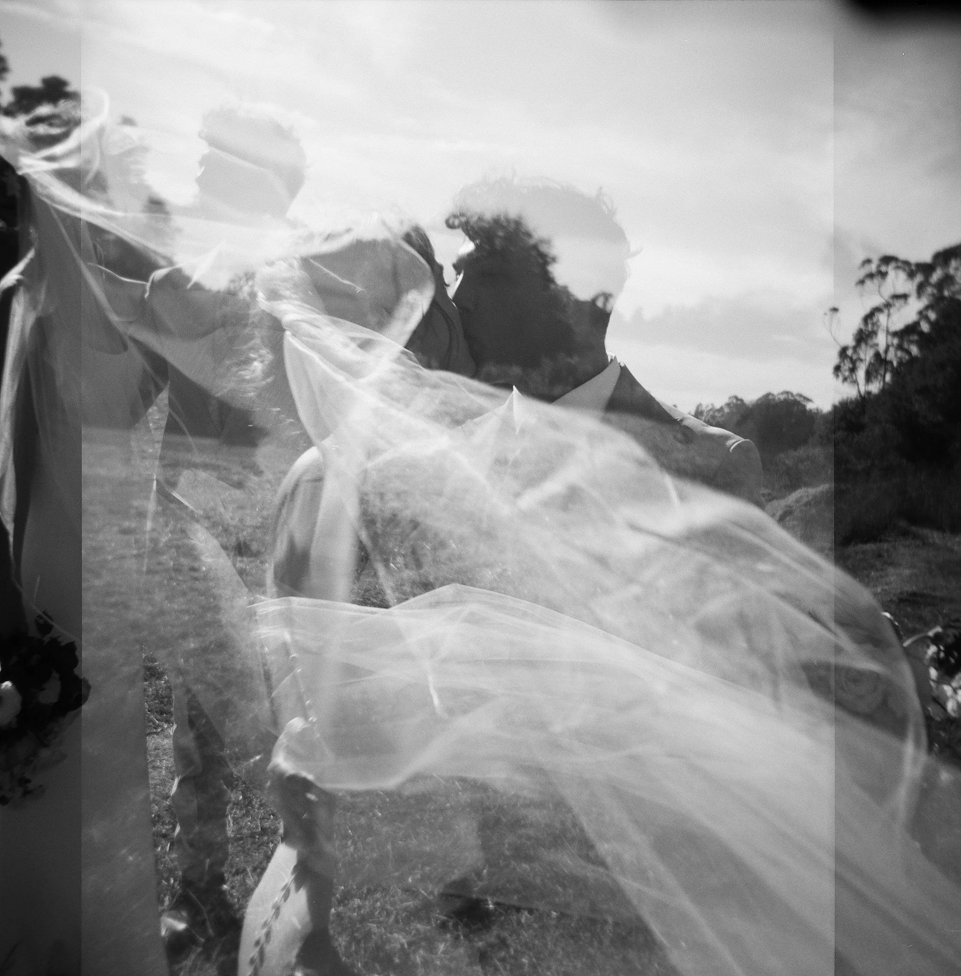 Black and white photo captured on film of a bride and groom kissing outdoors after their intimate wedding in Cambria, with trees in the background, their faces close together, and the bride wearing a veil.