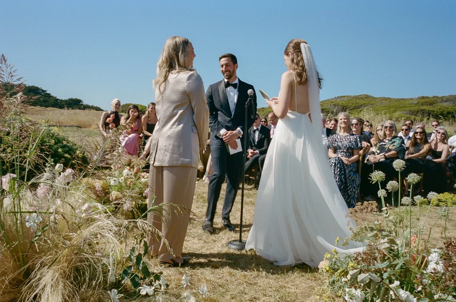 A couple getting married outdoors under a clear blue sky with guests seated behind them, with a officiant standing between them reading from a paper.