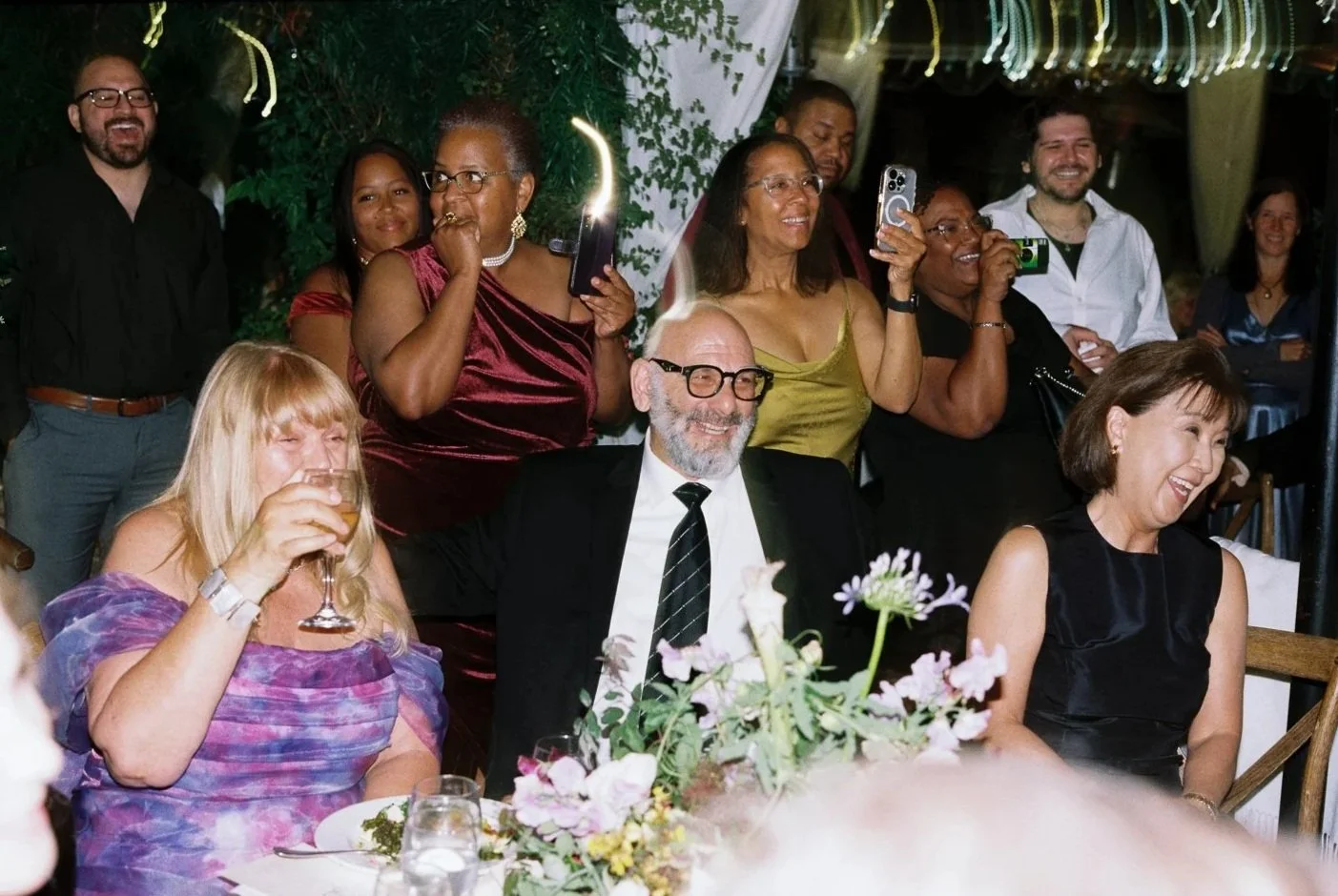 A group of people at a celebration, smiling and taking photos. Some are seated at a table with flowers, others are standing in the background.
