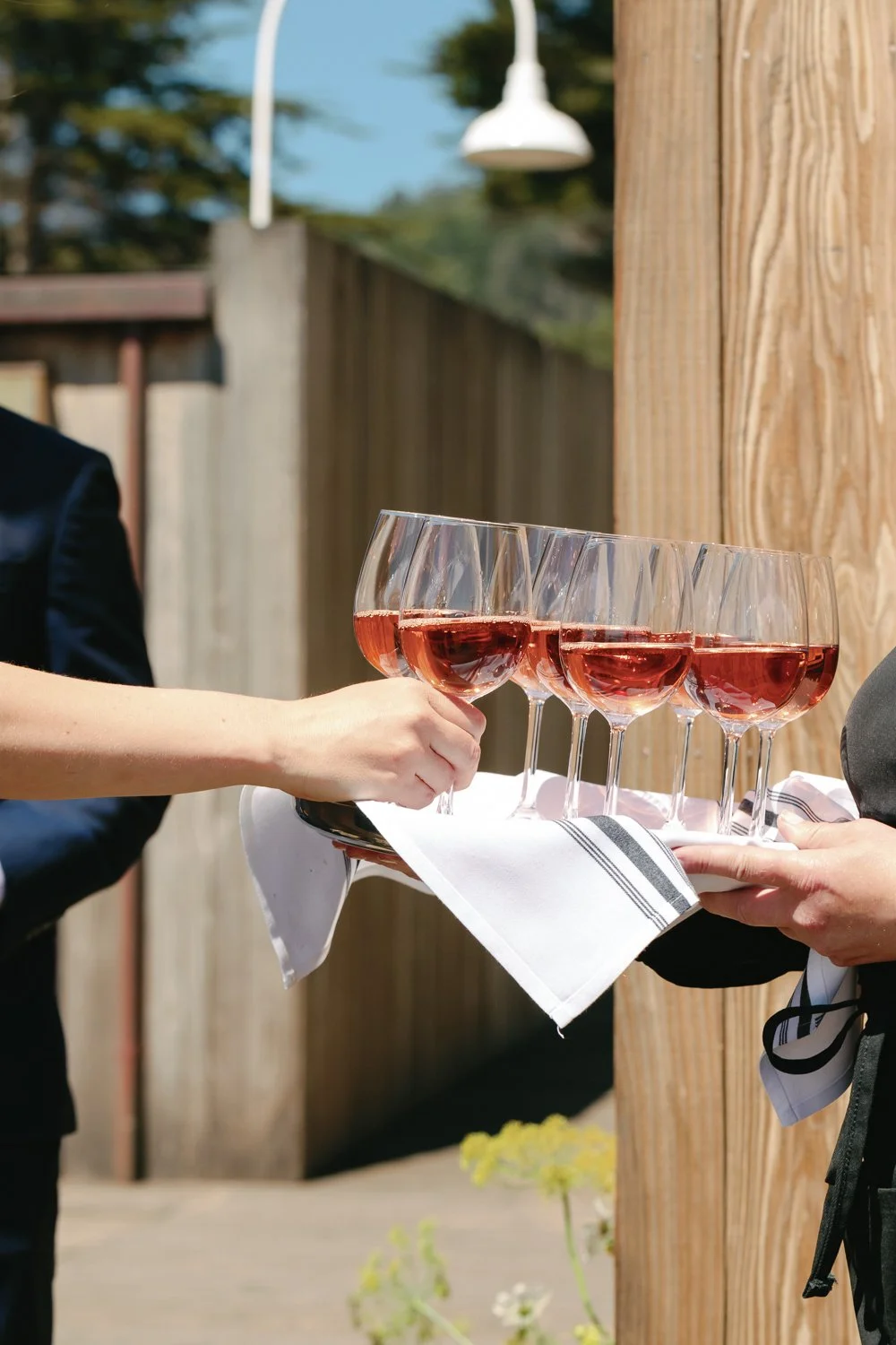 A person in a black suit presenting a tray of five glasses of rosé wine to another person during an outdoor event.