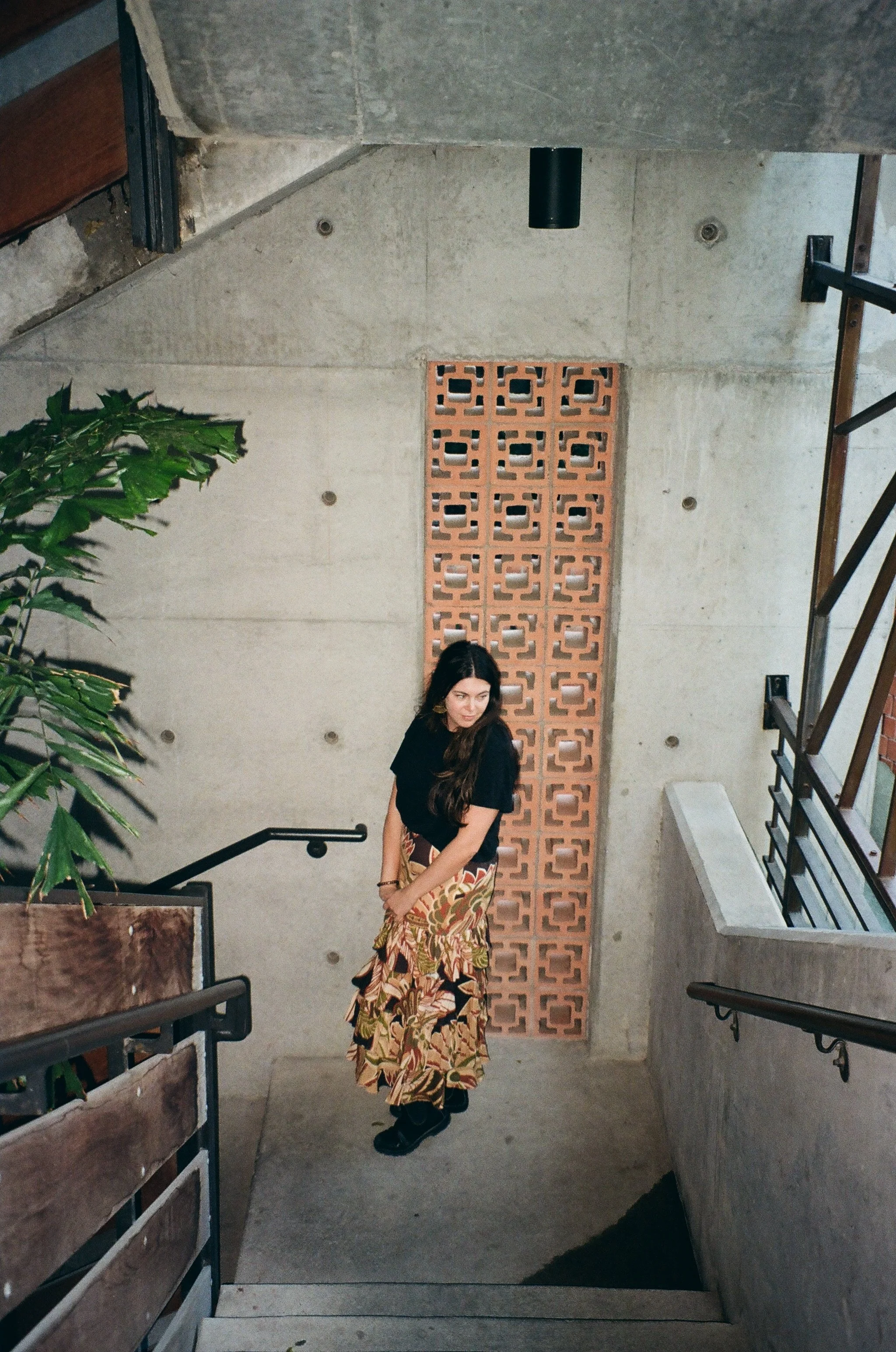 A woman with long hair wearing a black shirt and a colorful, patterned skirt standing on a staircase in an industrial-style interior with a concrete wall and a decorative brick partition behind her.