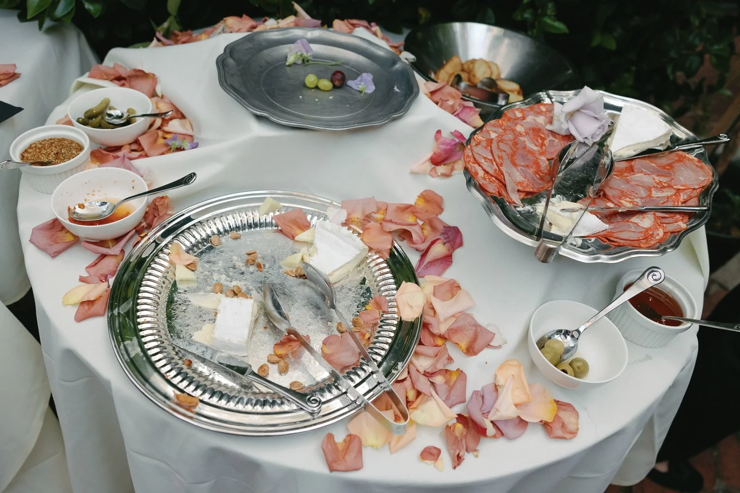 Empty dessert platter surrounded by rose petals and bowls of olives, sauces, and cured meats on a white tablecloth.