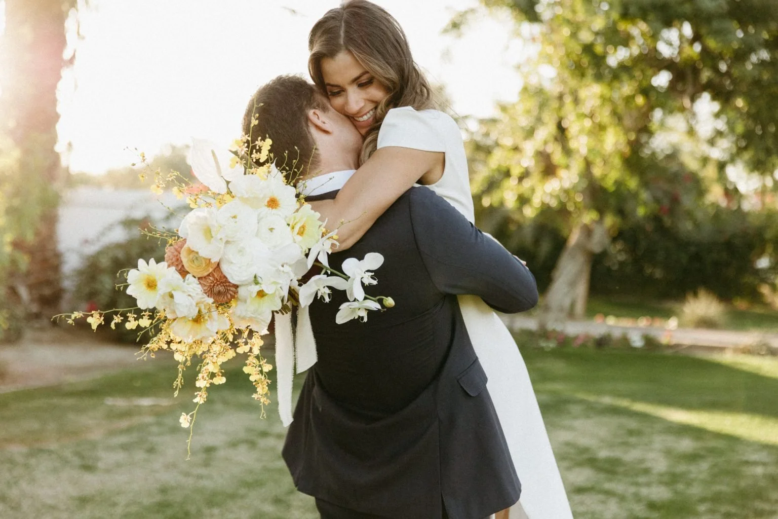 A woman in a white dress hugging a man in a black suit celebrating their marriage outside during sunset at the bougainvillea estate, holding a large bouquet of white and peach flowers, with trees and grass in the background.