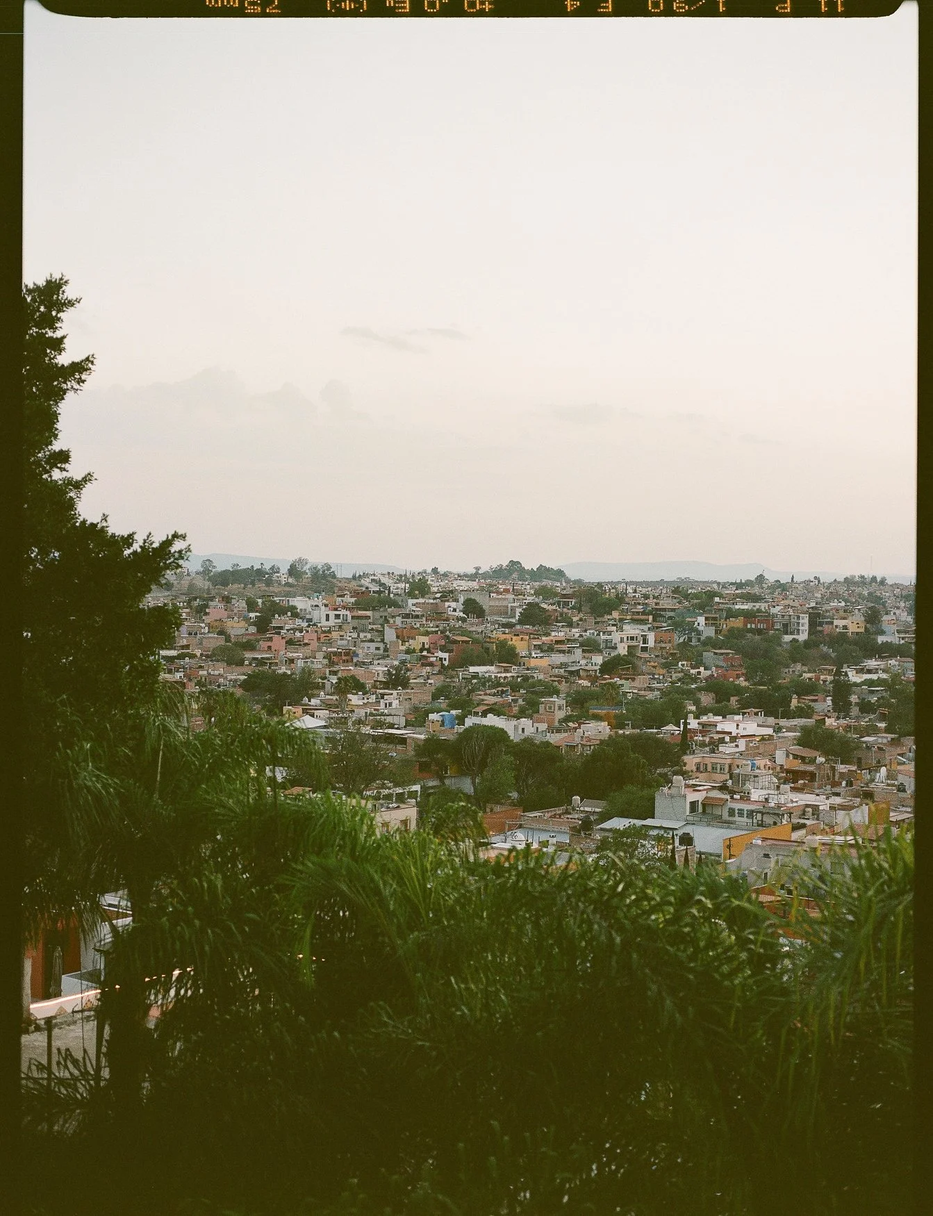 Hilly cityscape with dense low-rise homes, trees, and distant mountains under an overcast sky