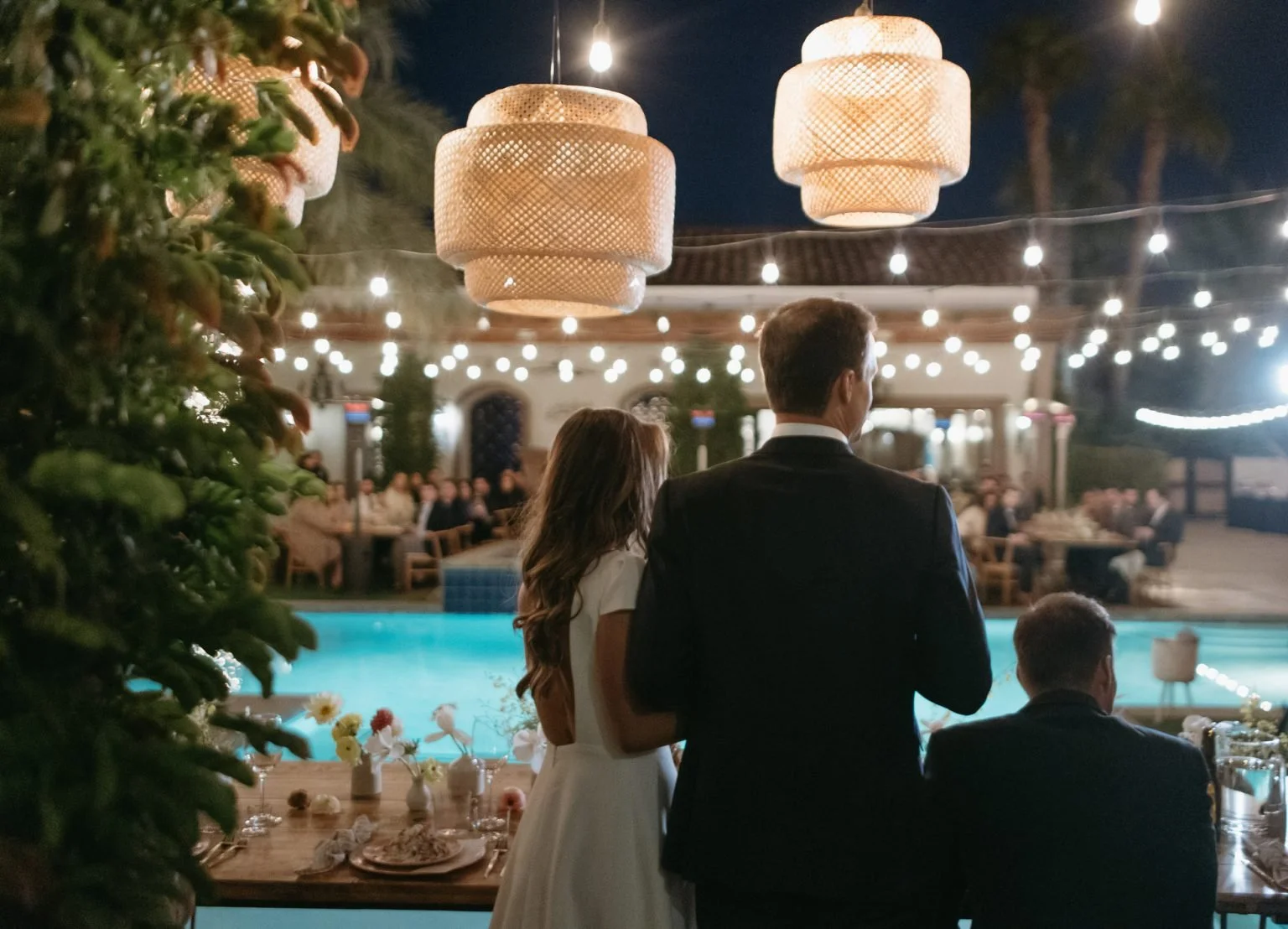 A couple at a nighttime outdoor event at the Bogainvillea estate, during their wedding reception, standing near a swimming pool with tables and guests in the background and hanging lights overhead.
