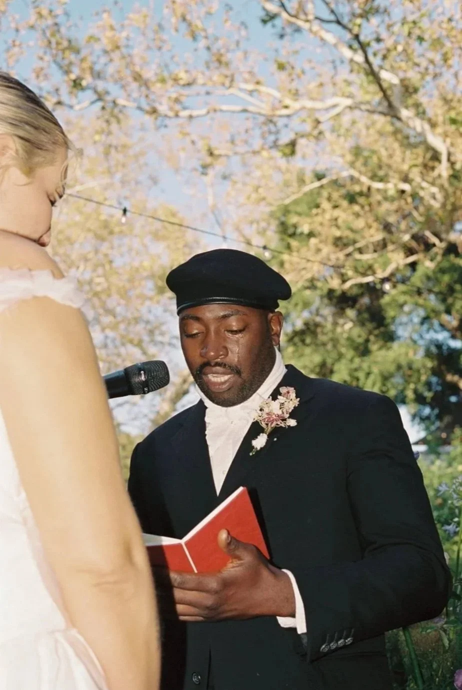 A man and woman at an outdoor wedding ceremony, with the man reading from a red book and the woman dressed in white, surrounded by trees with flowering branches.