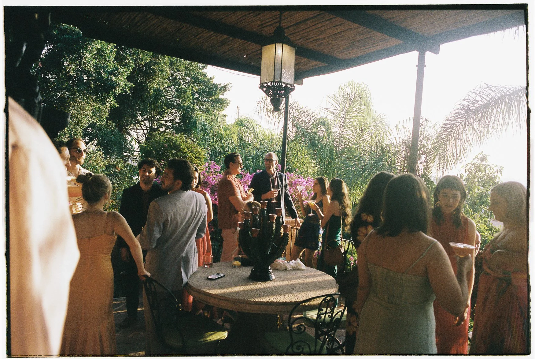 Guests mingle at an outdoor garden party on a covered patio surrounded by tropical plants, with drinks on a patio table.