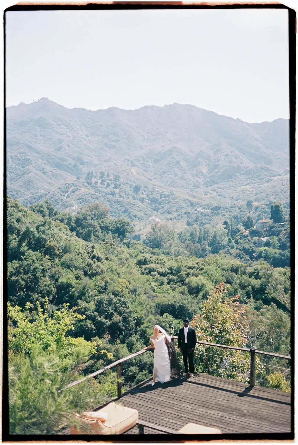 A bride and groom standing on a wooden deck overlooking a lush, green mountainous landscape.