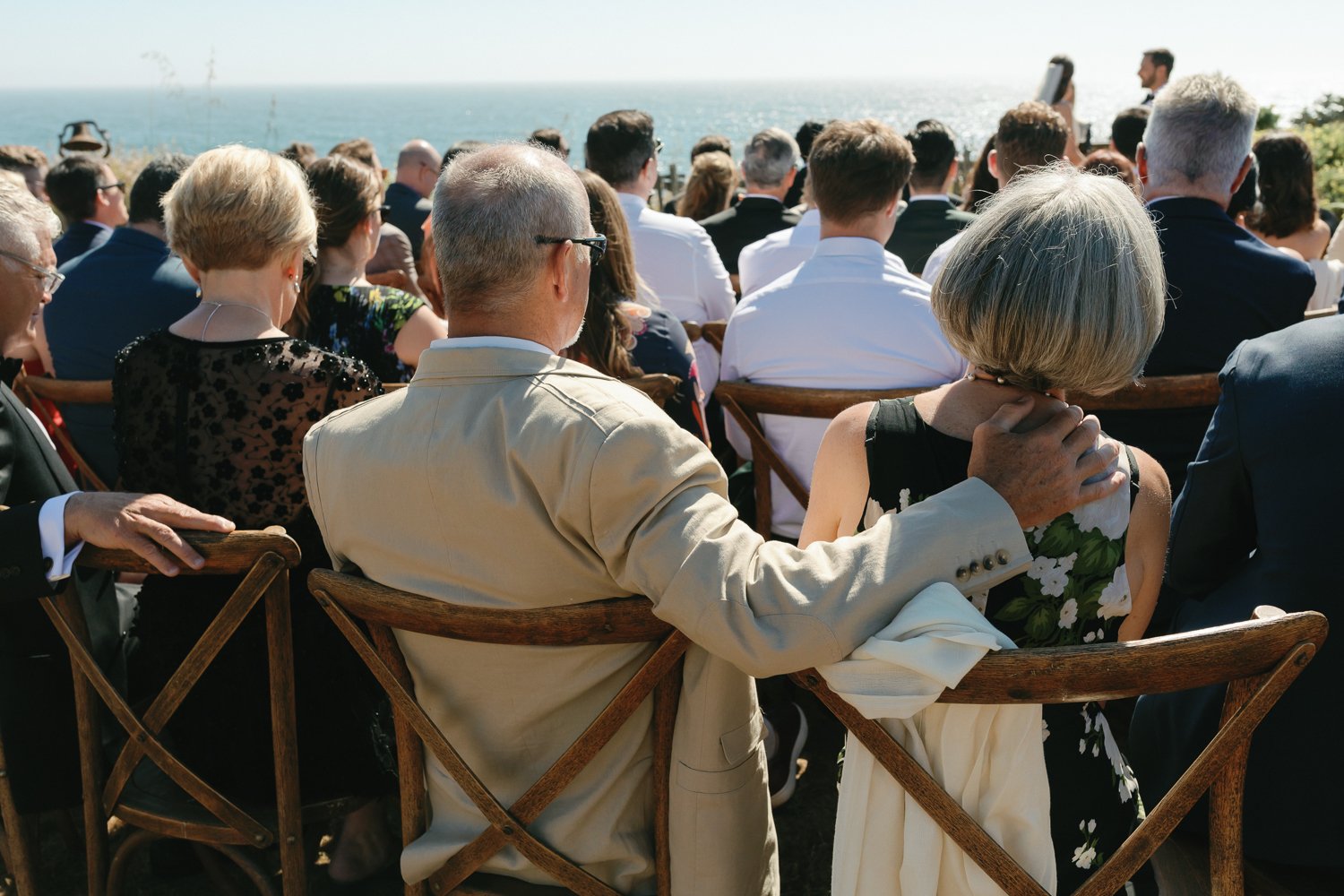 A group of formally dressed people sitting outdoors, facing a scenic ocean view with clear skies, attending a wedding or event during daytime.