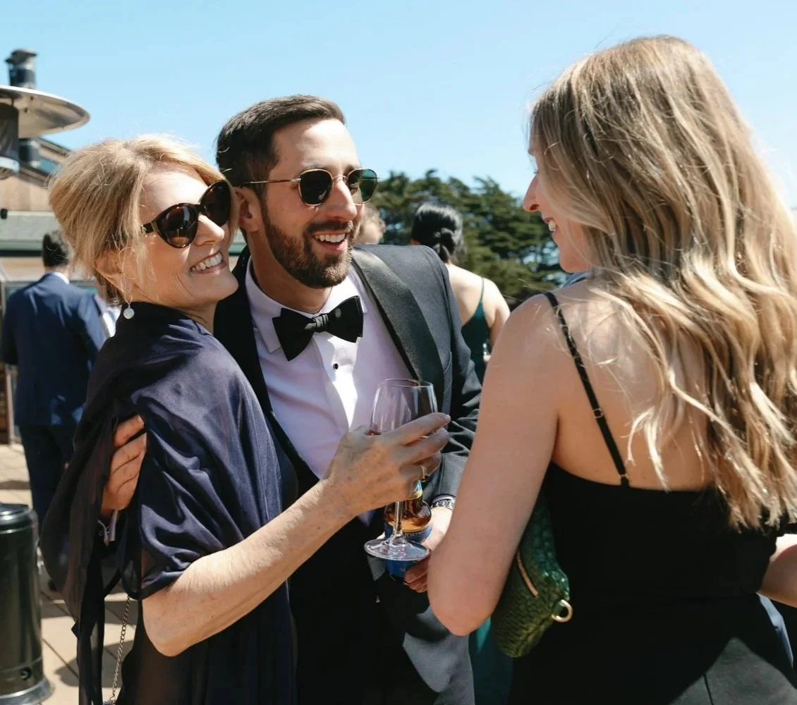 Three people in formal attire enjoying an outdoor event on a sunny day, with two women wearing black dresses and sunglasses and a man in a tuxedo with a bowtie and glasses, holding a glass of wine.