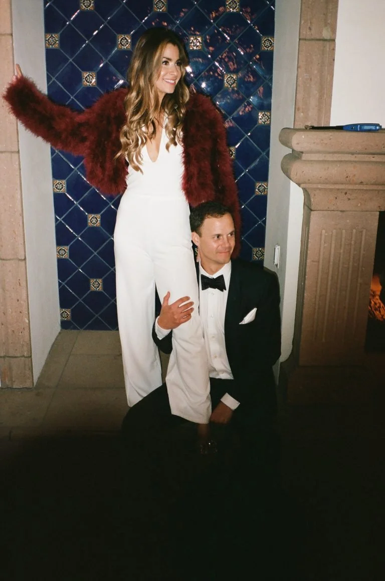 A woman in a white jumpsuit and a red furry jacket standing with her arm raised, smiling, beside a man in a black tuxedo and bowtie kneeling with one hand on his chest, in front of a blue tiled wall.