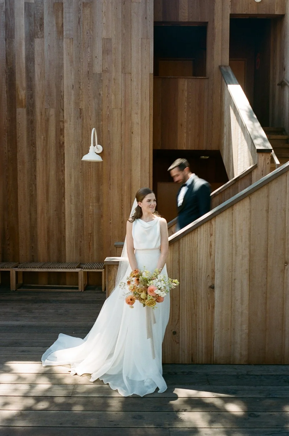 Bride holding a bouquet of flowers standing outdoors in front of a wooden wall with a groom blurred in the background.