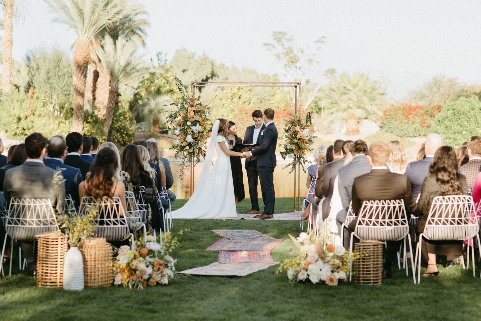 A wedding ceremony at the Boguainvillea estate overlooking a pond with a bride and groom standing in front of an officiant, outdoors surrounded by guests seated in rows, with floral arrangements and a decorative arch, in a lush garden setting.
