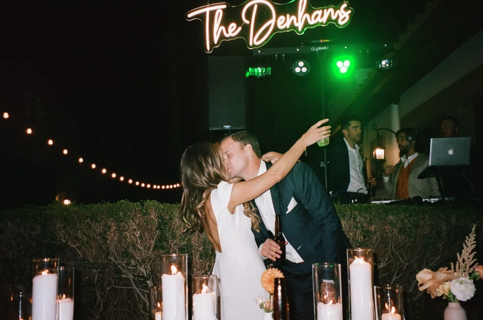 A couple kissing at a wedding reception, with candles on a table in the foreground and a DJ booth with people and green lighting in the background, and a neon sign that reads 'The Denhams'.