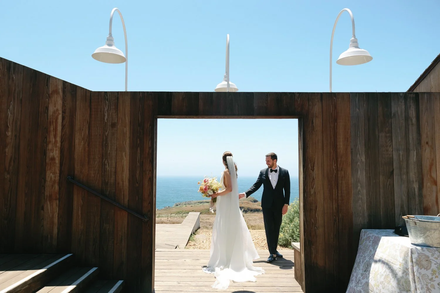 A bride and groom holding hands outside a wooden arch with ocean in the background on a sunny day.