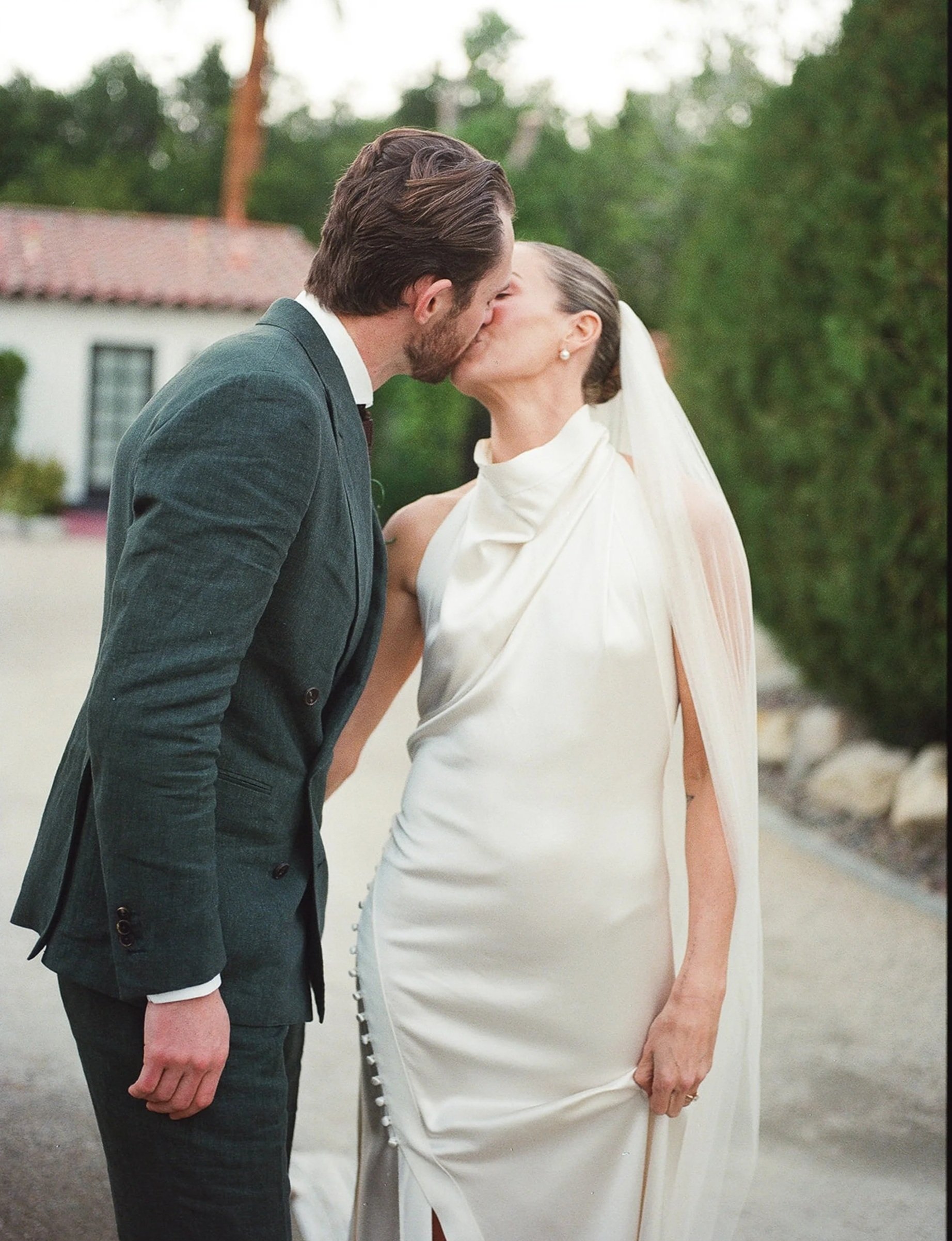 A man and woman are sharing a kiss outdoors at the Ingleside estate in palm springs wearing a danielle frankel dress with a the groom wearing a green linen suit.