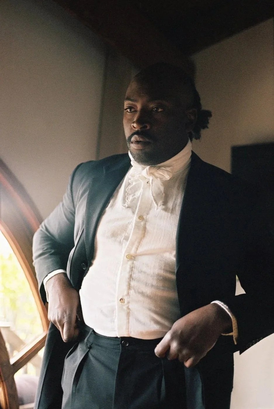 A man with dark skin wearing a black suit jacket and a white shirt with a ruffled neck, standing indoors near a window with natural light.