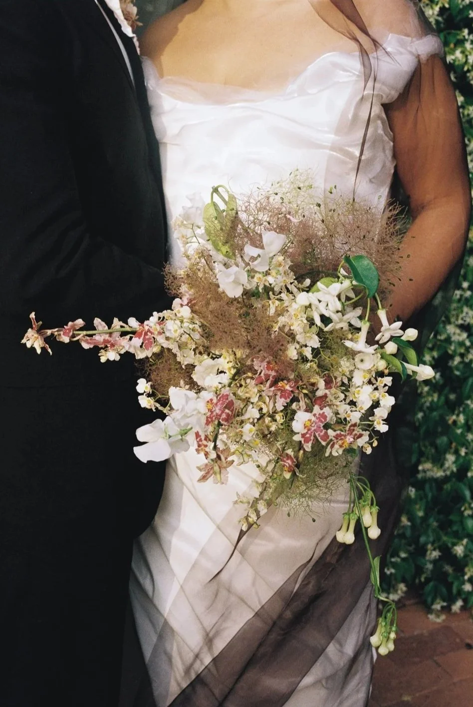 Bride in white wedding dress holding a floral bouquet  by Ou Vu floral studio with white and pink flowers, standing next to groom in black suit.