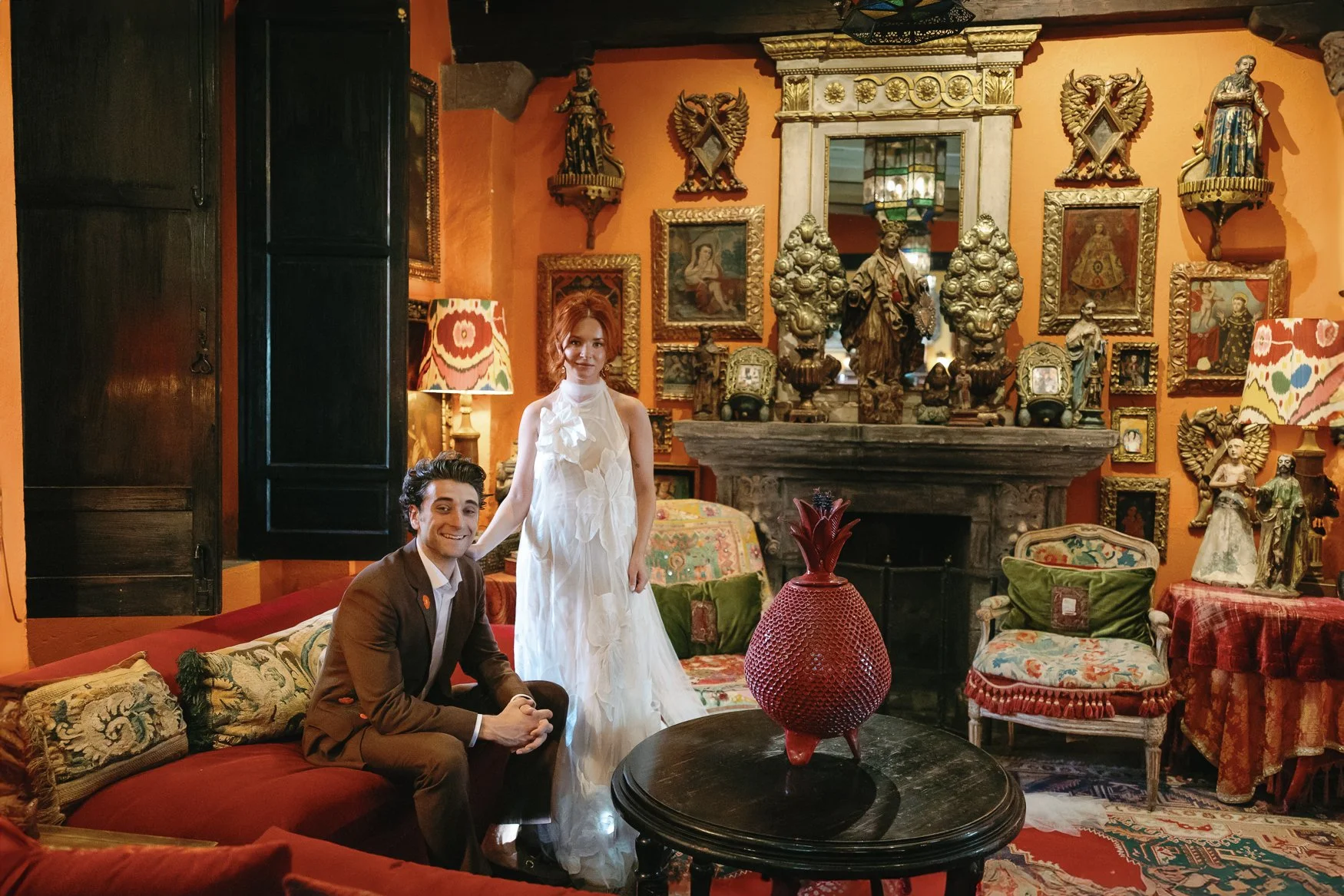 Couple posing in an ornate, art-filled living room with a fireplace, religious statues, framed artwork, and a red decorative vase at the unique wedding venue in san miguel de allende