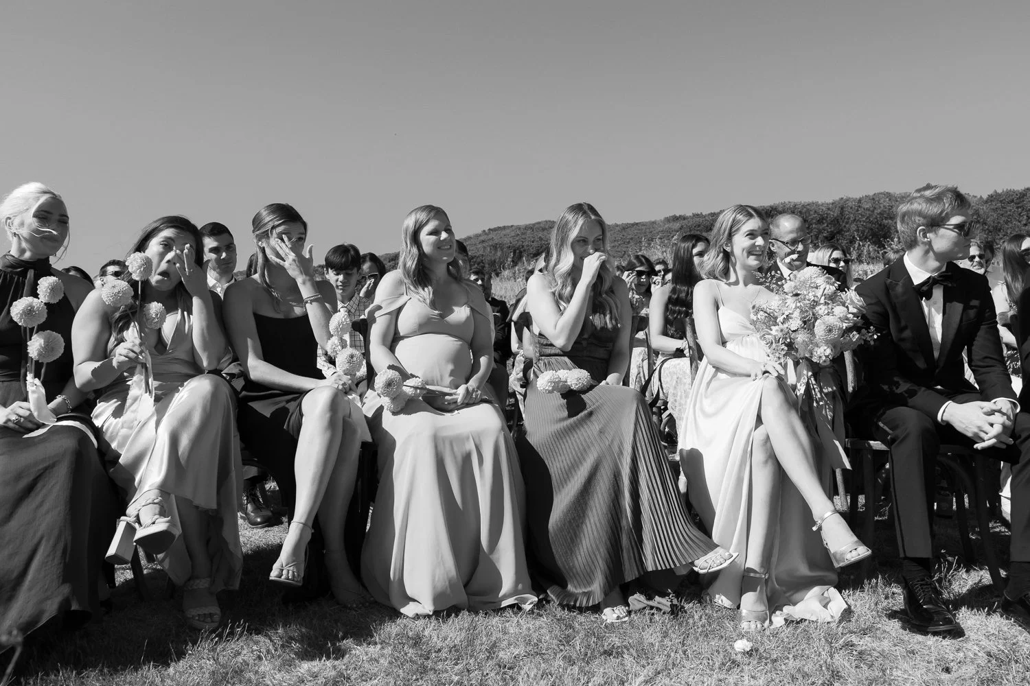 Black and white photo of a group of people attending an outdoor wedding ceremony, seated on chairs on grass, with a scenic backdrop of mountains and trees.