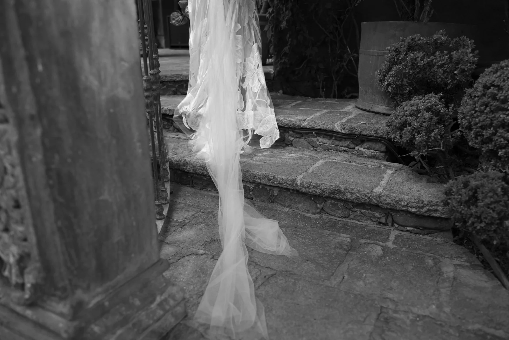 Black-and-white photo of a lace wedding dress train draped over stone steps in a garden