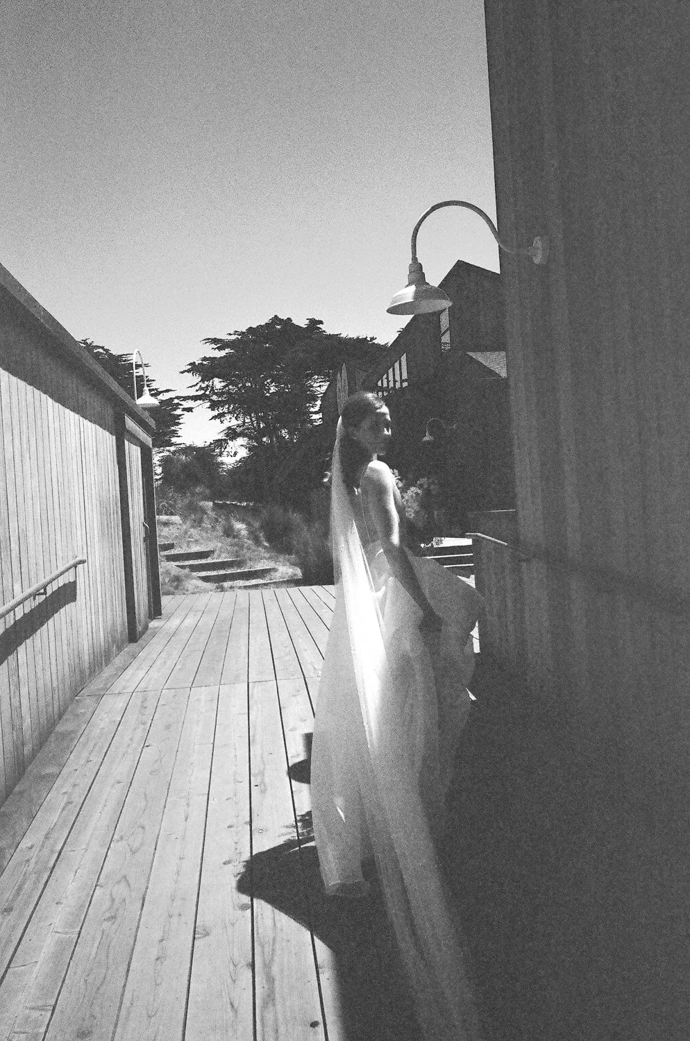 Black and white photo of a bride in a wedding dress with a veil standing on a wooden deck outside a house, illuminated by sunlight, with trees and a clear sky in the background.