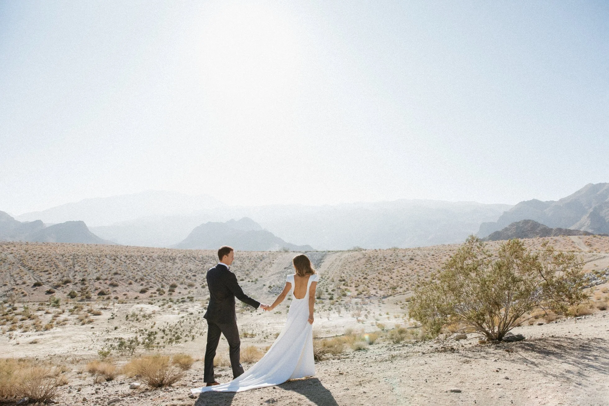 A couple in wedding attire holding hands with the Palm Springs desert landscape with mountains in the background.