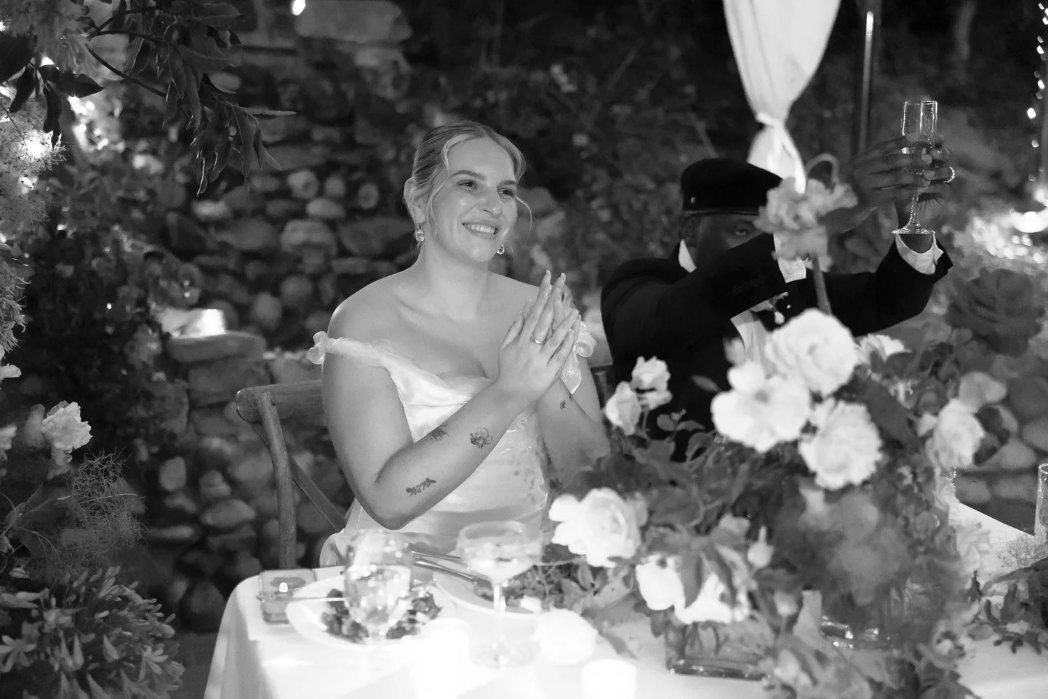 A smiling woman in a wedding dress sitting at a table decorated with flowers, with a man in a suit and hat raising a glass in a toast, at an outdoor evening wedding celebration.