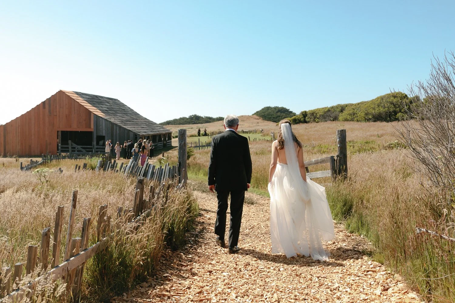A bride and a groom walking along a dirt path towards a rustic barn for their outdoor wedding ceremony, with guests gathered near the barn.