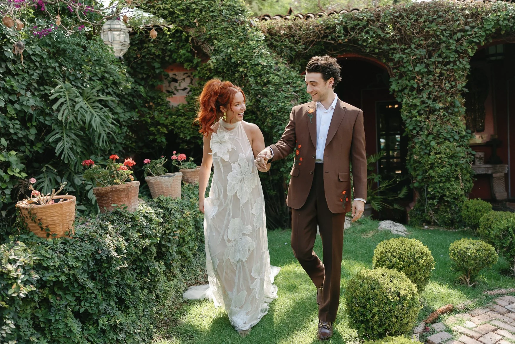 Newlywed couple holding hands and walking through a lush garden, wearing a floral wedding dress and brown suit