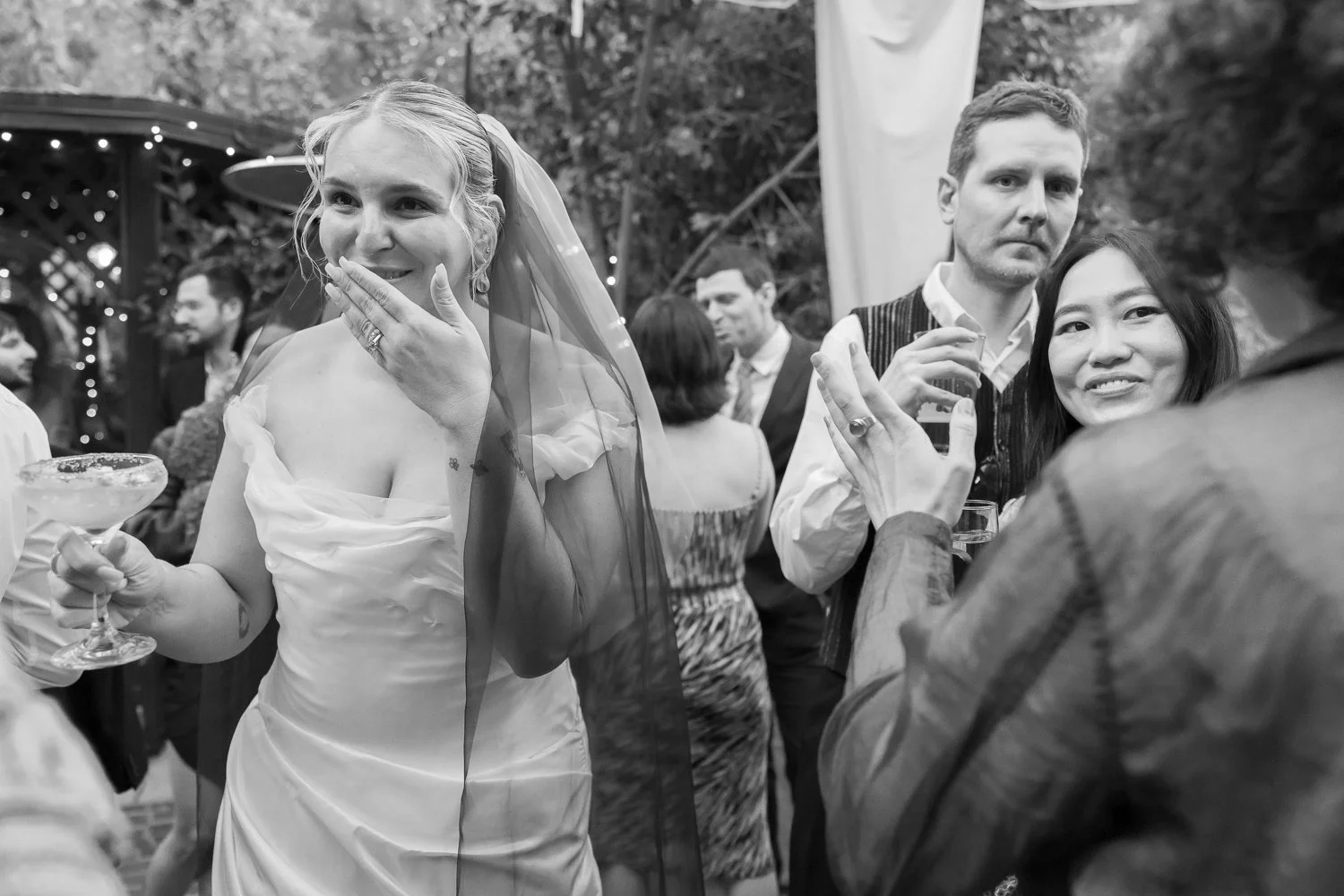 Black and white photo of a woman in a wedding dress holding a cocktail, smiling with her hand covering her mouth, at a celebration with other people dancing and talking in the background.