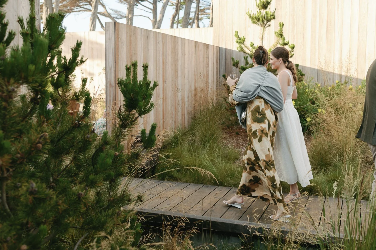 Two women in formal attire walking on a wooden pathway in a garden, with one holding a glass and the other smiling, surrounded by greenery and a wooden fence.