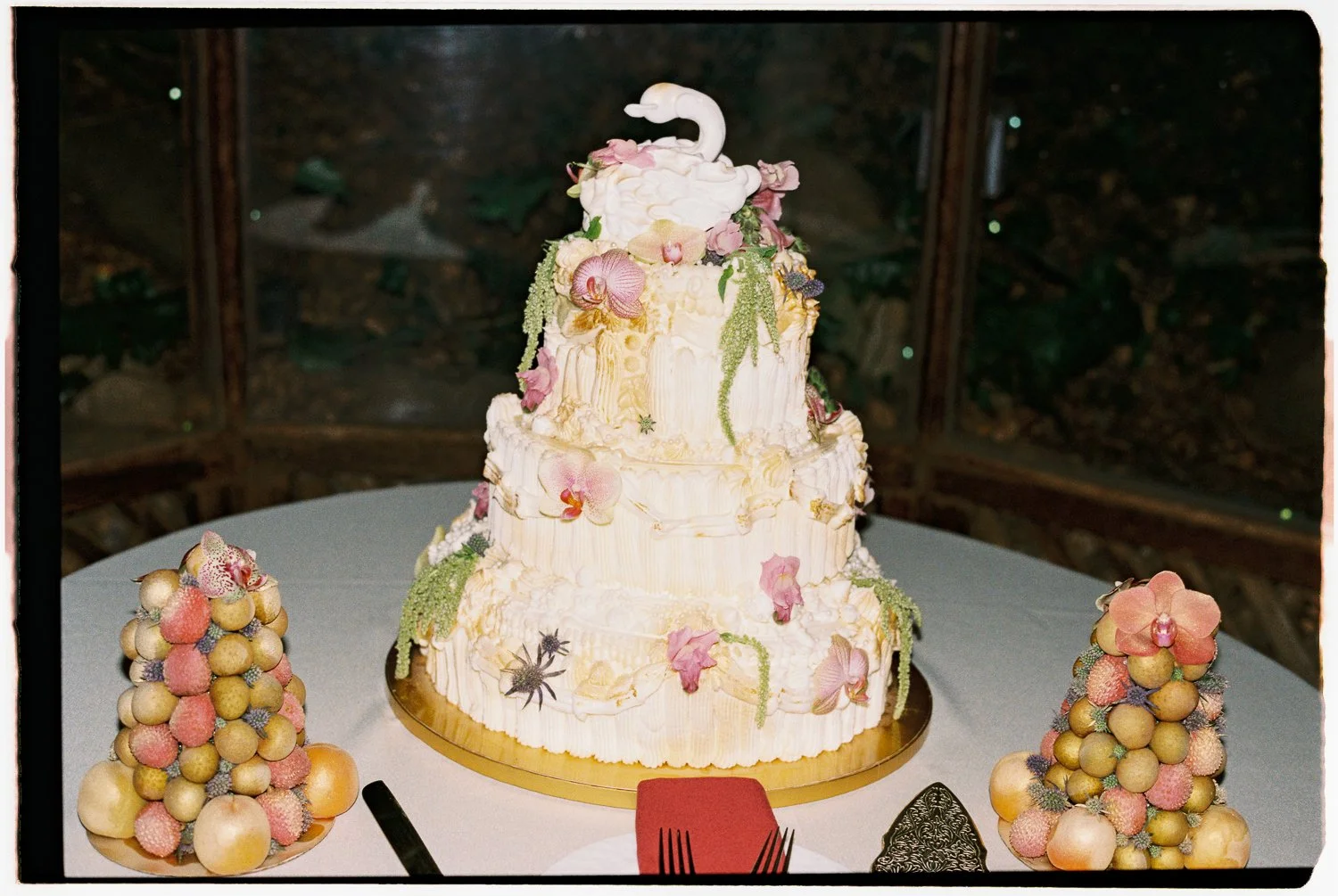 A multi-tiered wedding cake decorated with edible flowers and a  hand made swan topper, surrounded by two cone-shaped arrangements of small fruits on a round table.