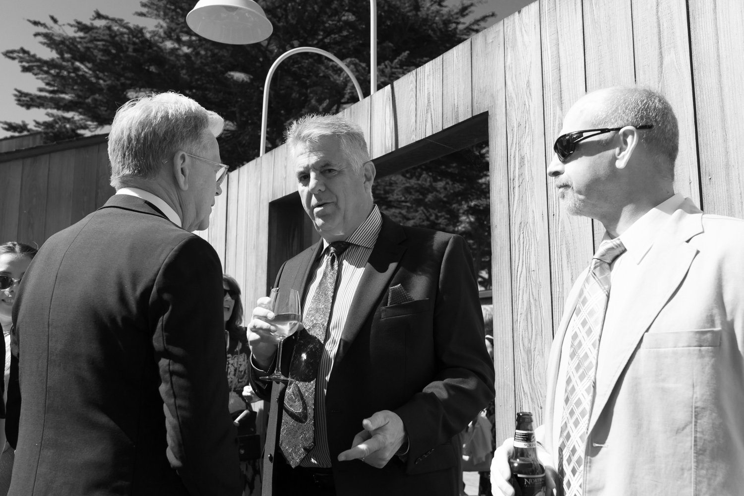 Three men in suits and ties talking outdoors during a social event, with one man holding a wine glass and another holding a beer bottle, near a wooden fence.