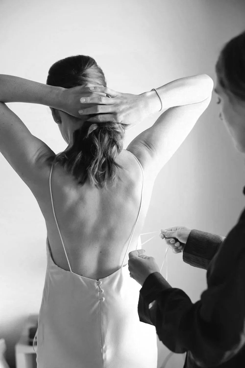 A woman in a white backless dress is having a medical procedure, possibly a tattoo, with a person holding medical equipment near her lower back. She has her hands behind her head, and her hair is tied back.