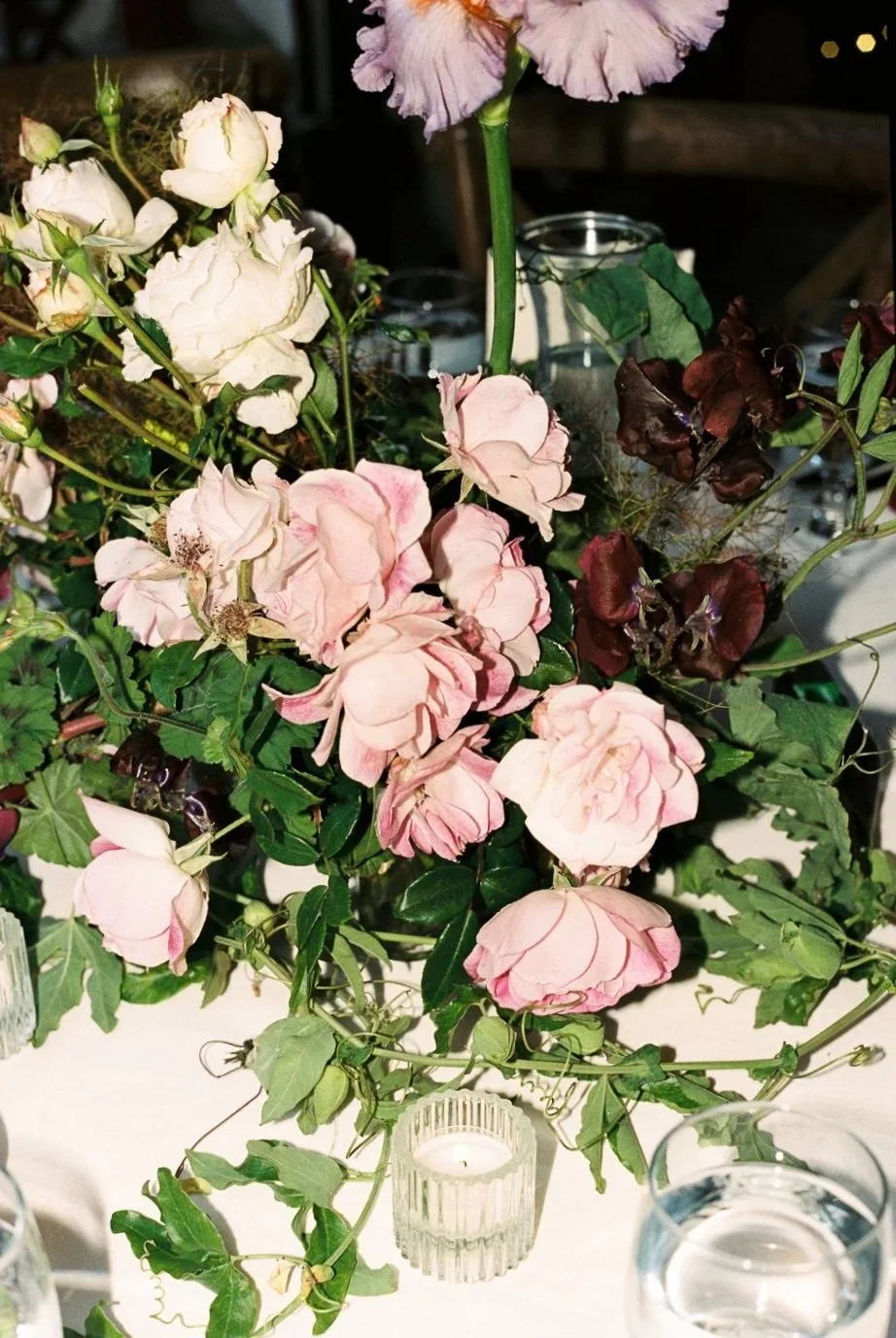 A floral arrangement featuring pink, white, and purple flowers, with candles and greenery on a table.
