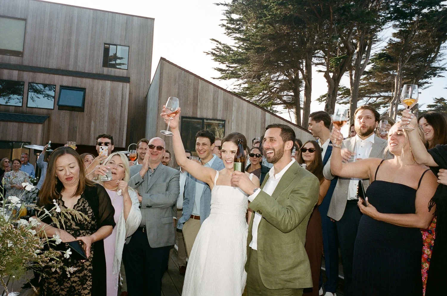 Group of people at an outdoor celebration, toasting with drinks, smiling, and enjoying the event near a modern building and trees.