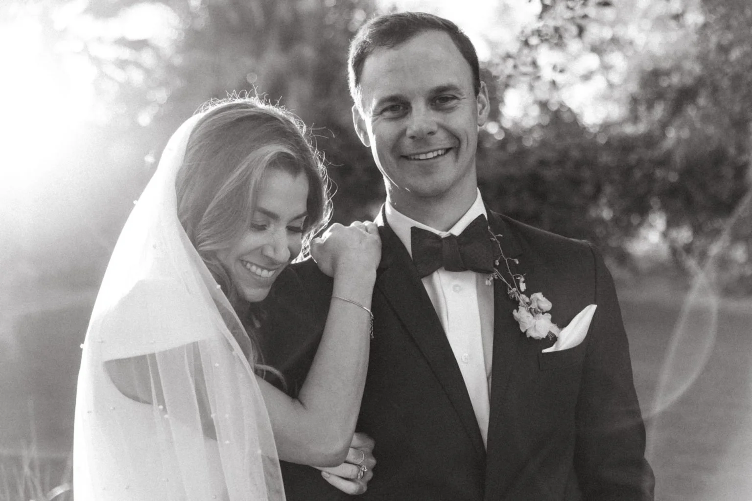 A black and white photo of a wedding couple outdoors after their wedding, with the bride leaning on the groom's shoulder and smiling down, and the groom smiling directly at the camera, dressed in formal wedding attire with a bow tie and boutonniere.