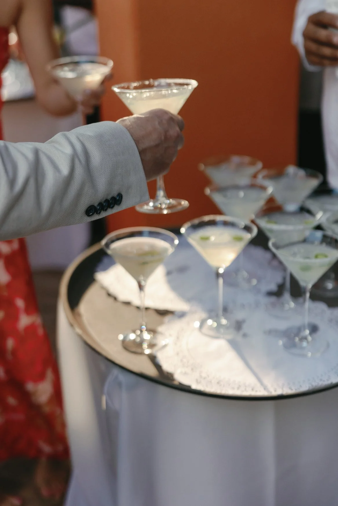 Person in a white jacket holding a pale cocktail at a cocktail reception with several martini glasses on a tray